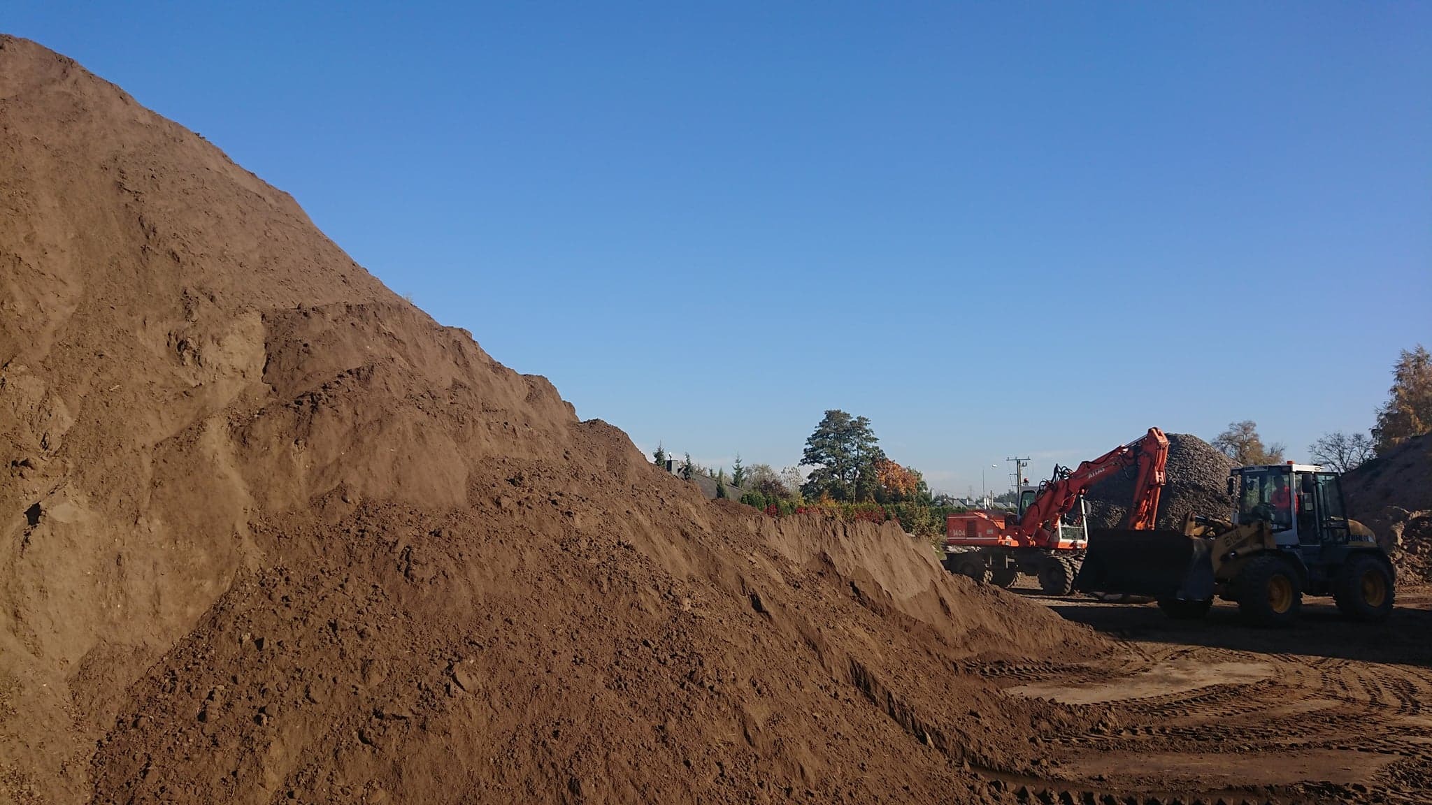 Góra ciemnej ziemi z widocznymi śladami opon i pracującymi w tle koparką i ładowarką, błękitne niebo.