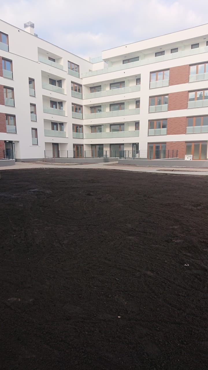 Świeżo rozplantowana czarna ziemia ogrodowa na dziedzińcu nowego apartamentowca z białymi elewacjami i przeszklonymi balkonami.