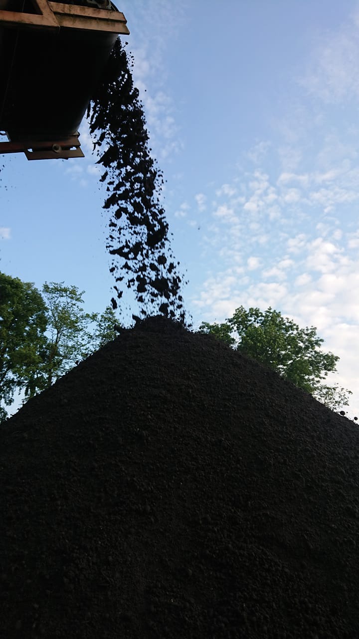 Proces wysypywania ciemnej ziemi z metalowego pojemnika na dużą pryzmę, błękitne niebo z chmurami w tle.