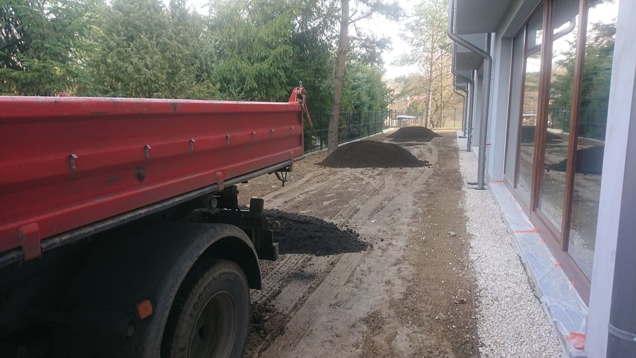 Czerwona wywrotka częściowo wysypuje materiał na posesji w Warszawie; w tle widoczne pryzmy materiału oraz budynek z dużymi oknami.