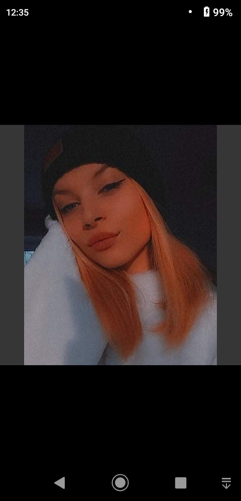 Portret młodej kobiety z blond włosami i czarną czapką, ubrana w biały sweter, z delikatnym makijażem oczu i ust, pozująca w zacienionym otoczeniu.