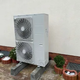 Biała, dwuwentylatorowa jednostka zewnętrzna na betonowych wspornikach, ustawiona przy jasnej ścianie budynku obok czerwonych donic z krzewami i resztkami śniegu.