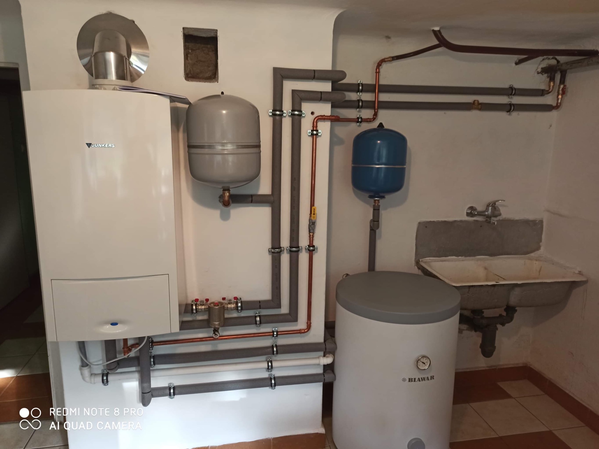 Biały piec Junkers z podłączeniami, szary zbiornik wyrównawczy, niebieski zbiornik ciśnieniowy i biały zasobnik Biowar w kotłowni z umywalką.