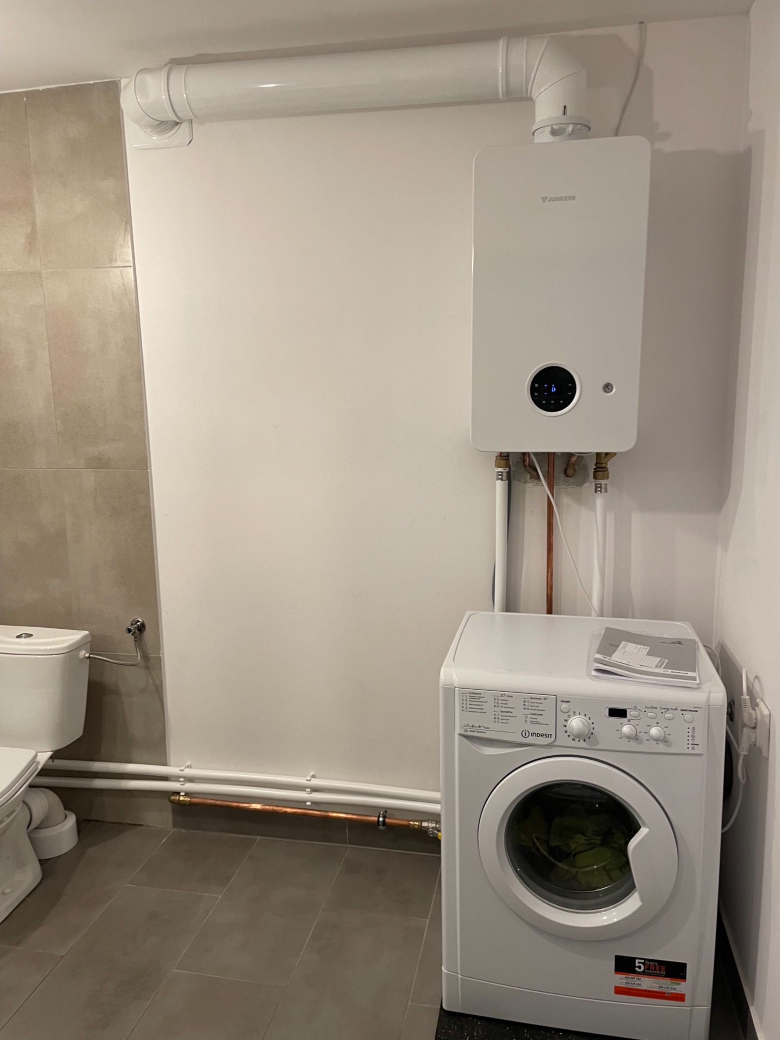 Kompaktowy, wiszący piec grzewczy Junkers z odprowadzeniem spalin, zainstalowany w łazience nad pralką Indesit, z widocznymi rurami miedzianymi i elementami instalacji.