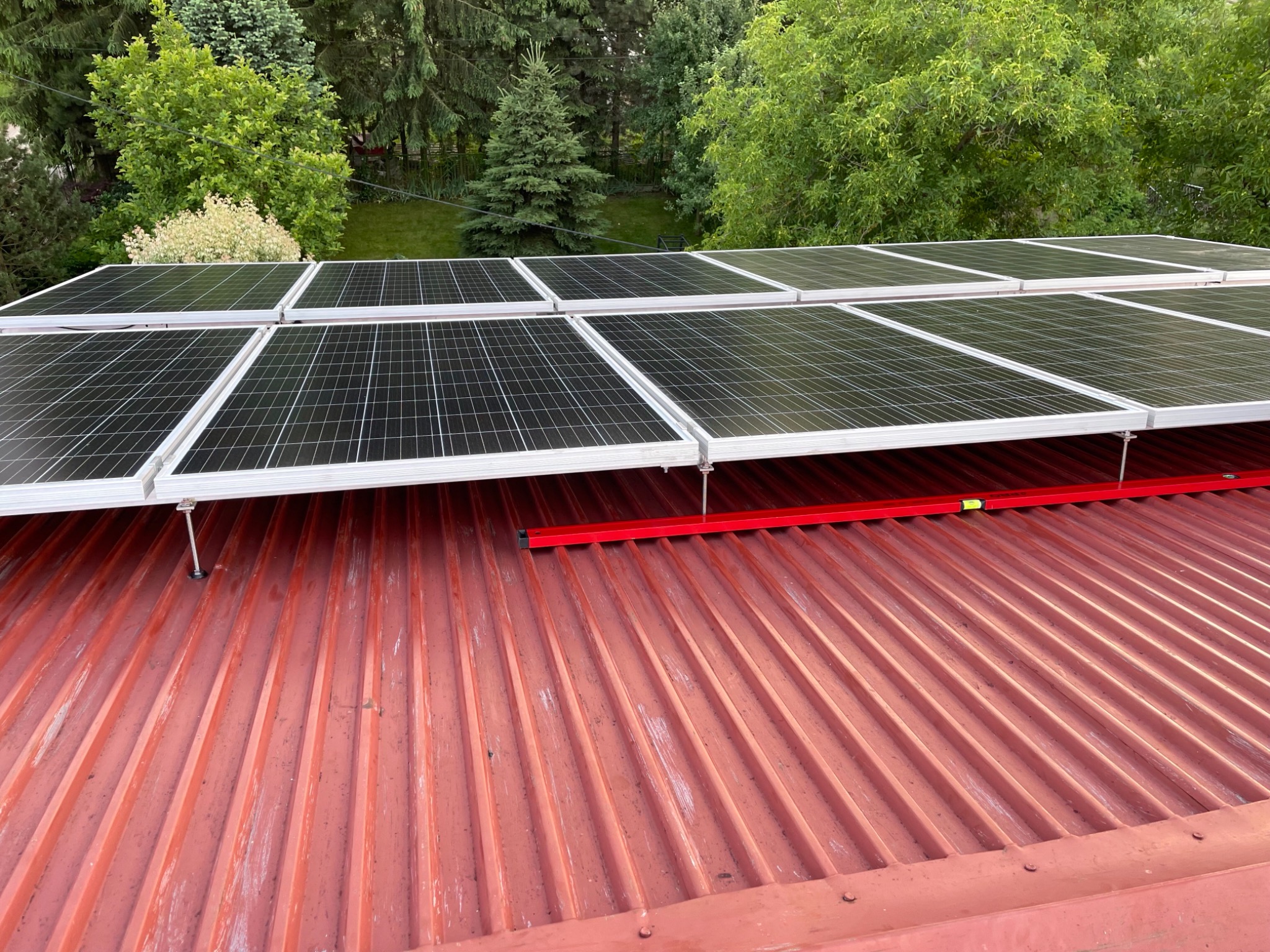 Montaż paneli fotowoltaicznych na blaszanym dachu z użyciem poziomicy w słoneczny dzień, widok z góry na tle zieleni.