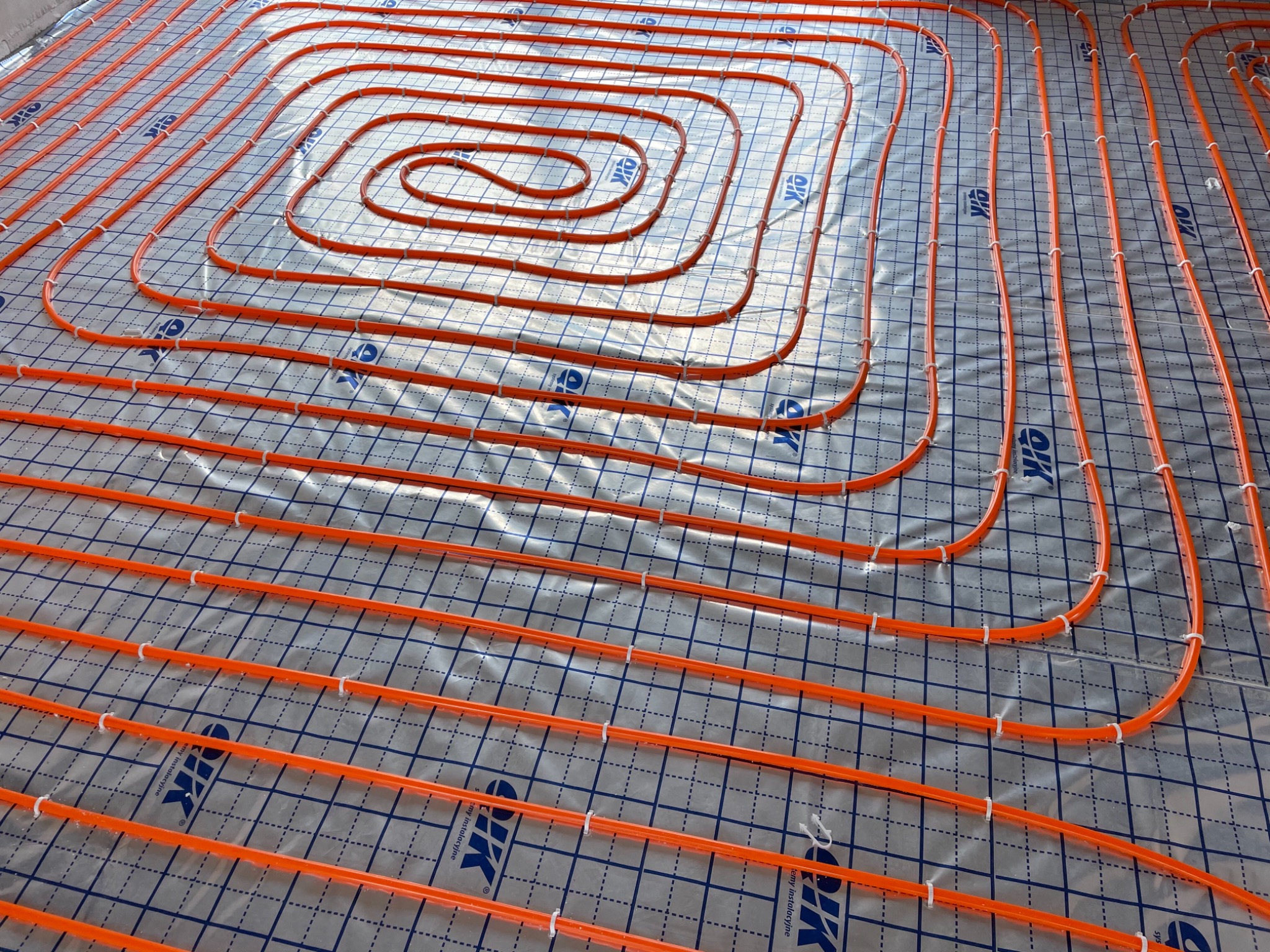 Pomarańczowe rury ogrzewania podłogowego ułożone spiralnie na srebrnej folii z niebieską siatką, przymocowane białymi klipsami.