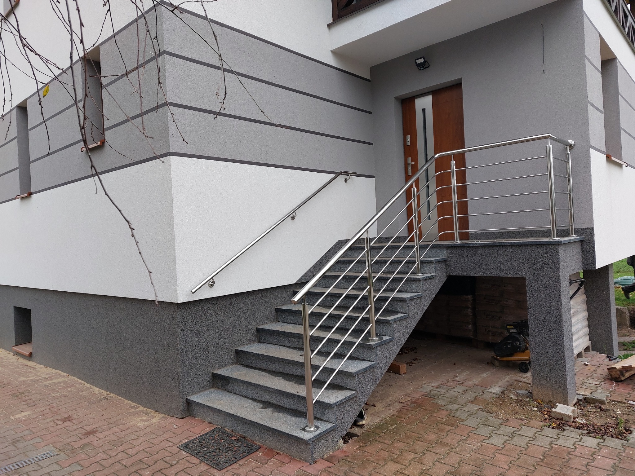 Zewnętrzne schody z szarych płyt z nierdzewną balustradą i poręczą, prowadzące do wejścia w nowoczesnym domu z elewacją w odcieniach szarości i bieli.