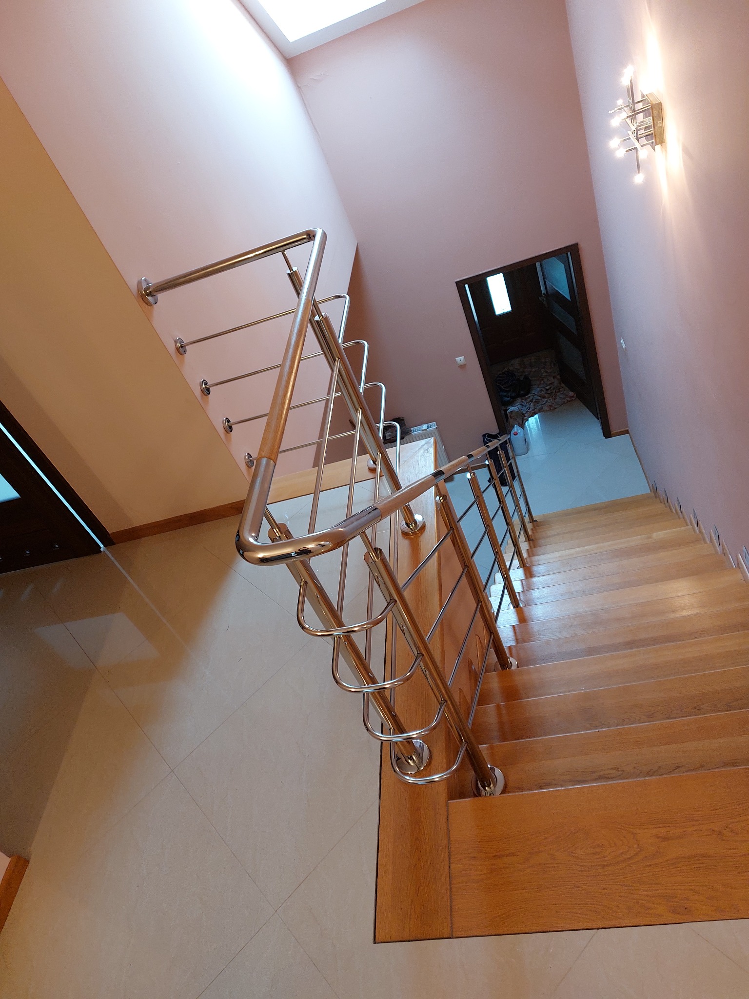 Wnętrze domu z drewnianymi schodami i metalową balustradą z drewnianym elementem, widziane z góry, z widocznym oknem dachowym i nowoczesną lampą ścienną.