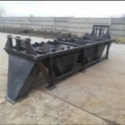 Czarna, metalowa konstrukcja spawana, prawdopodobnie element maszyny przemysłowej, na zewnątrz, szary beton i płot w tle.