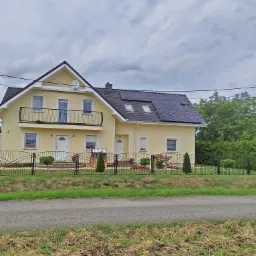 Żółty dom jednorodzinny z czarnym dachem i panelami fotowoltaicznymi, widoczny zza metalowego ogrodzenia.