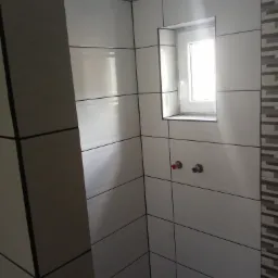 Ściany w łazience wyłożone białymi płytkami z ciemnymi fugami, widoczne wyprowadzenia instalacji wodnej i okno wnękowe, dekoracyjna ściana z płytek mozaikowych po prawej stronie.