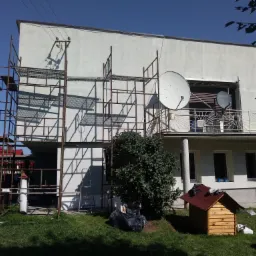 Dom jednorodzinny w trakcie malowania elewacji, widoczne rusztowania, satelity na balkonie i psia buda na trawniku.