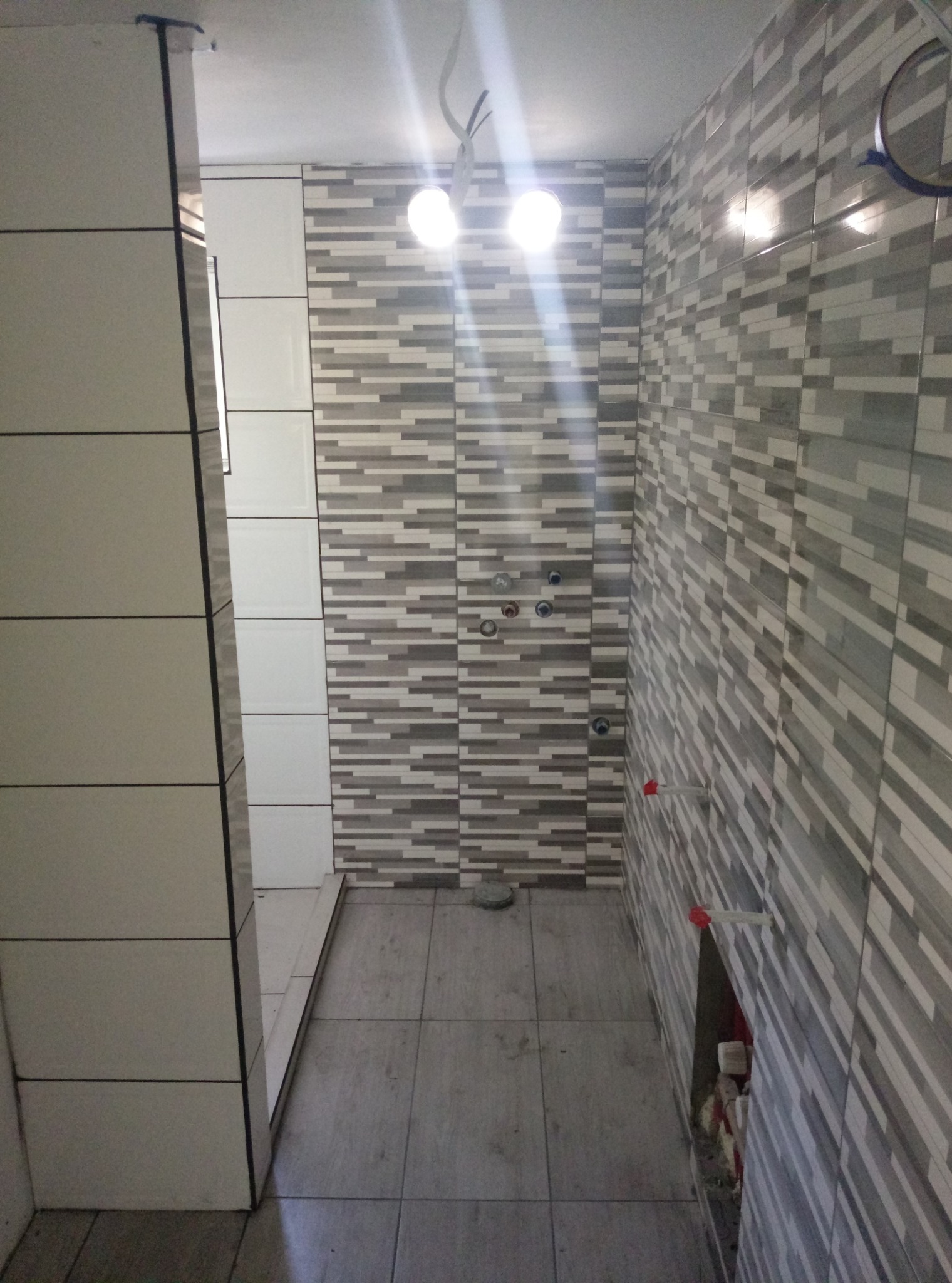 Wnętrze łazienki w trakcie prac wykończeniowych; widoczne ułożone płytki w odcieniach szarości i bieli na ścianach, przygotowane instalacje wodne oraz częściowo wykończony prysznic z białymi płytkami.