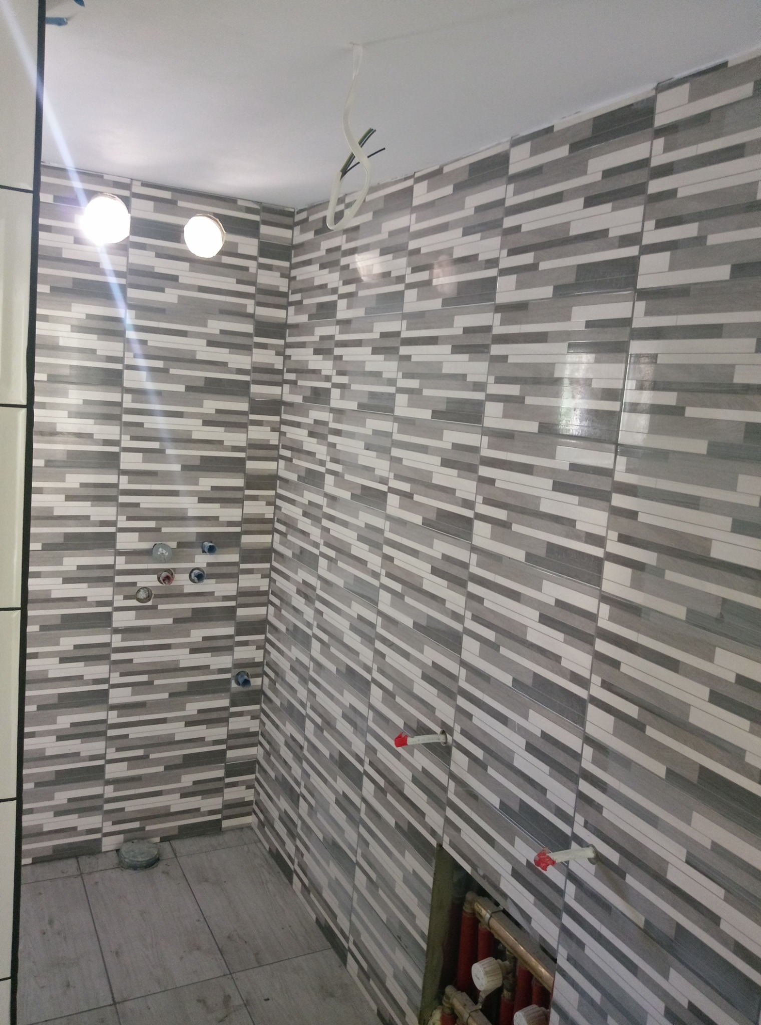 Wnętrze łazienki w trakcie prac wykończeniowych, widoczne ułożone płytki w odcieniach szarości, przygotowane punkty instalacji wodnej i fragment instalacji grzewczej w dolnym rogu.