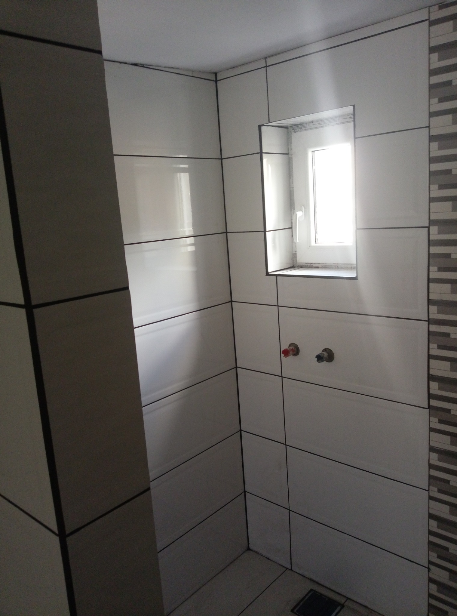 Ściany w łazience wyłożone białymi płytkami z ciemnymi fugami, widoczne wyprowadzenia instalacji wodnej i okno wnękowe, dekoracyjna ściana z płytek mozaikowych po prawej stronie.