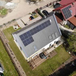 Widok z drona na nowoczesny dom z szarym dachem, na którym zamontowane są panele słoneczne. Na dachu widoczne okna dachowe. Obok domu znajduje się drewniany taras z meblami ogrodowymi i zadbany...