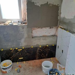 USŁUGI REMONTOWO BUDOWLANE KAROL GRABAREK - Łazienka w trakcie remontu: częściowo położone ciemne płytki z żółtymi krzyżykami dystansowymi, ściany w surowym tynku, widoczne narzędzia i materiały budowlane.