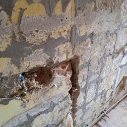 USŁUGI REMONTOWO BUDOWLANE KAROL GRABAREK - Ściana w trakcie skuwania tynku, z odsłoniętymi instalacjami hydraulicznymi i fragmentami cegieł. Widoczne ślady po starych warstwach farby i zaprawy.
