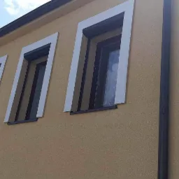 Żółta elewacja budynku z trzema oknami z roletami, obramowanymi białymi listwami dekoracyjnymi, widok z dołu.