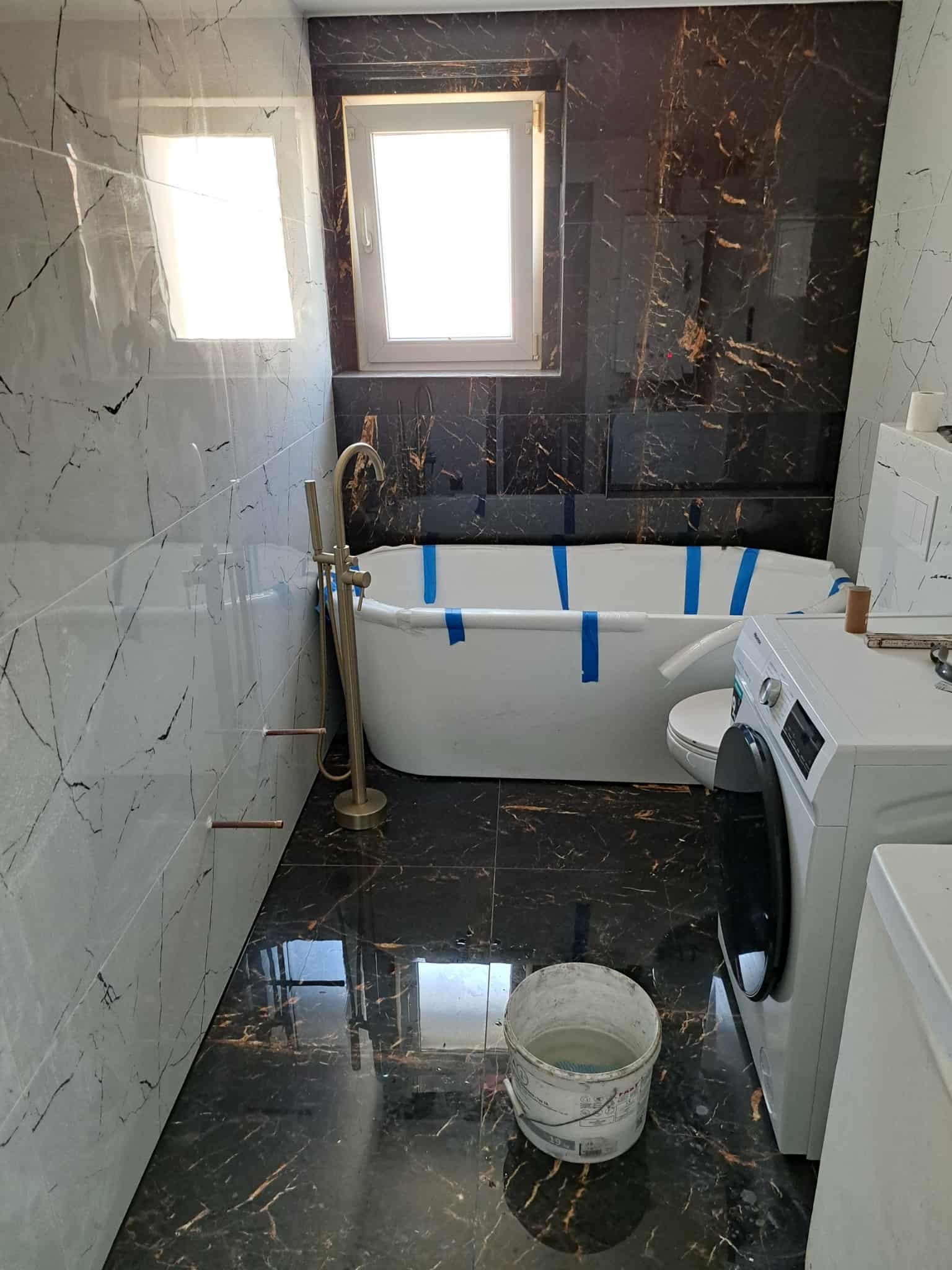 Wykończona łazienka z czarno-białymi płytkami imitującymi marmur, wolnostojąca wanna zabezpieczona taśmą, złota armatura i pralka. Widoczny kubeł.