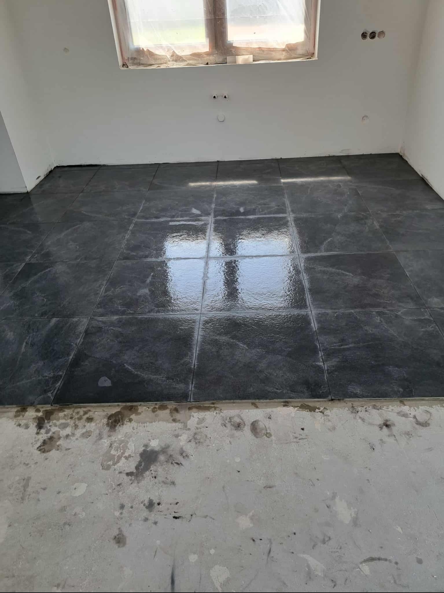 Ułożone, ciemne płytki podłogowe z marmurkowym wzorem w jasnym pomieszczeniu. Widoczne ślady po pracach na podłodze przy ścianie. Okno zabezpieczone folią.