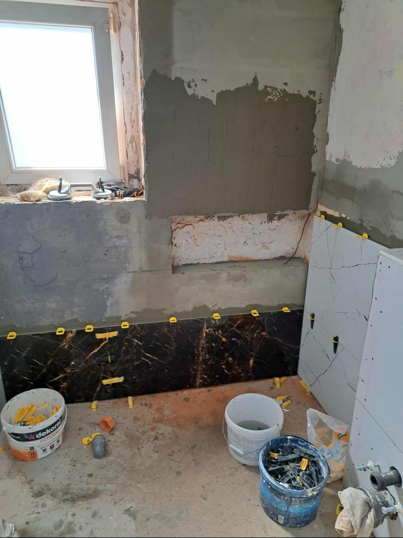 Łazienka w trakcie remontu: częściowo położone ciemne płytki z żółtymi krzyżykami dystansowymi, ściany w surowym tynku, widoczne narzędzia i materiały budowlane.