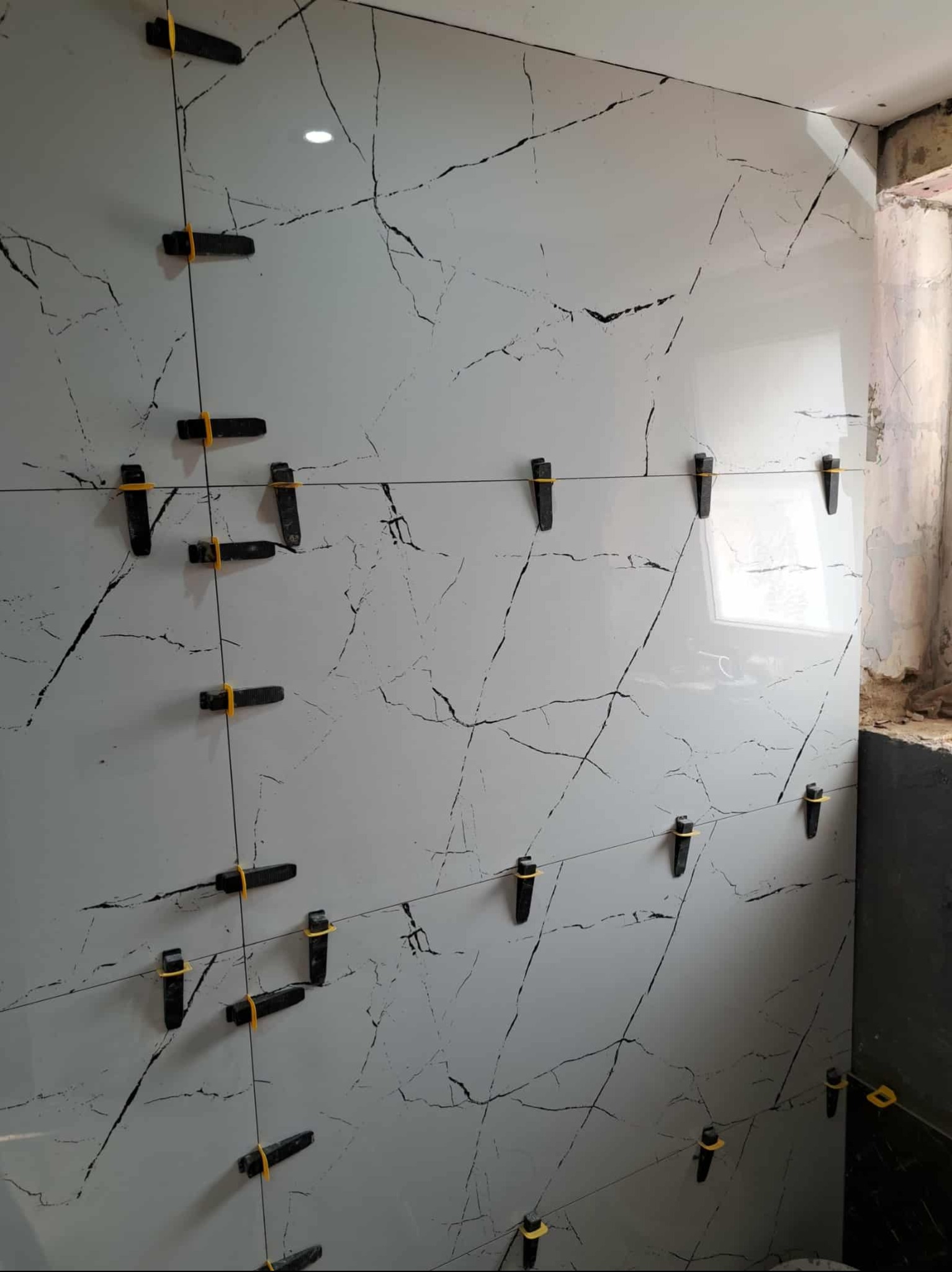 Ściana wyłożona białymi płytkami z czarnym wzorem żyłkowania, z użyciem klipsów poziomujących, w trakcie prac wykończeniowych w łazience.