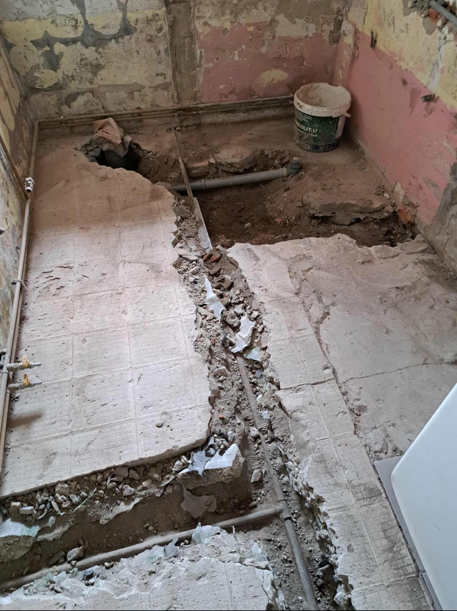 Demontaż starej podłogi w łazience z odsłoniętymi rurami. Widoczne gruz i resztki płytek. Ściany z resztkami farby. Etap prac przygotowawczych do remontu.