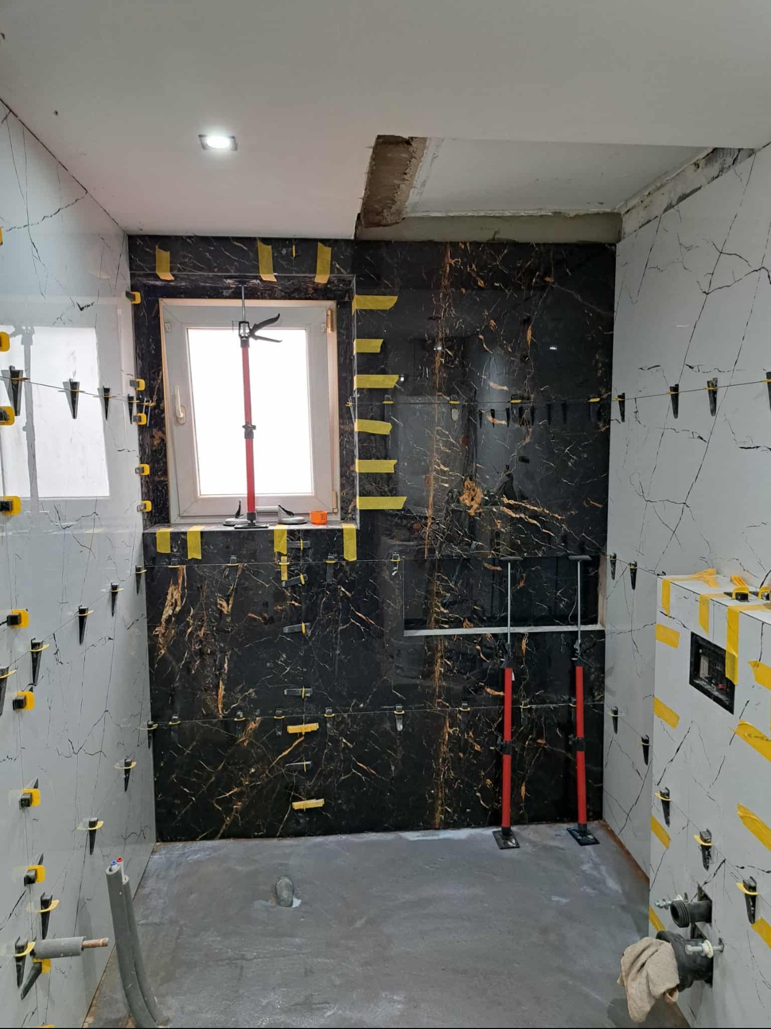 Łazienka w trakcie prac glazurniczych: czarne i szare płytki imitujące marmur, system poziomowania płytek, podpory teleskopowe. Widoczne okno i fragment instalacji.