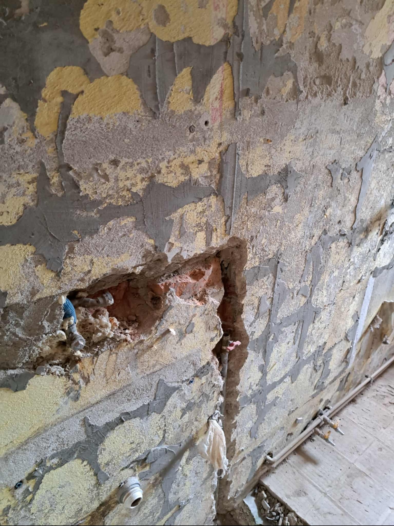 Ściana w trakcie skuwania tynku, z odsłoniętymi instalacjami hydraulicznymi i fragmentami cegieł. Widoczne ślady po starych warstwach farby i zaprawy.