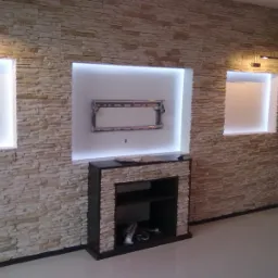 Ściana z dekoracyjnej cegły z podświetlanymi wnękami, jedna z ramą pod telewizor, poniżej czarny kominek z półkami, widoczne przewody wewnątrz.