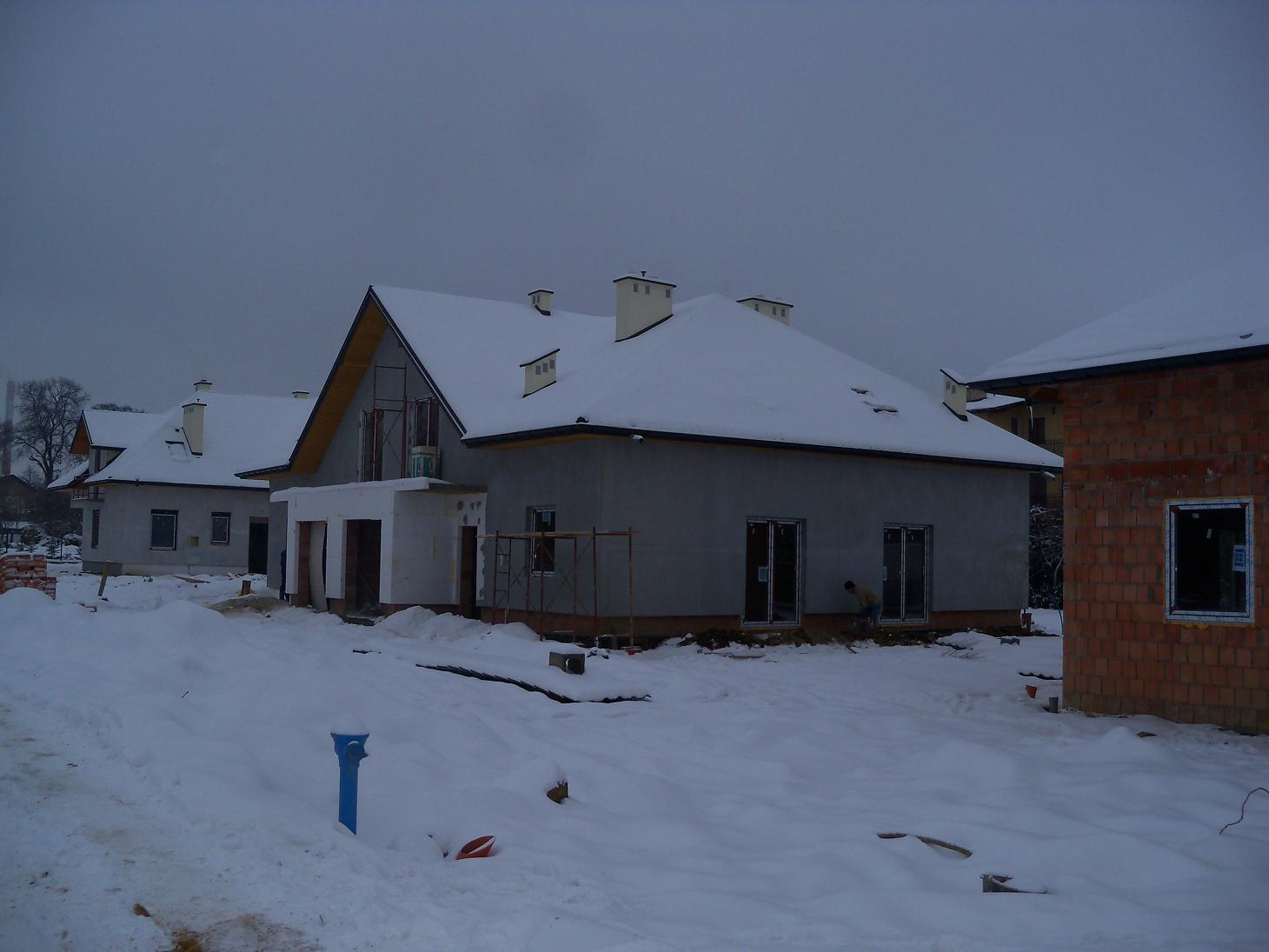 Domy w stanie surowym zamkniętym, częściowo ośnieżone, widoczne rusztowania, teren budowy pokryty śniegiem, pochmurne niebo w tle.