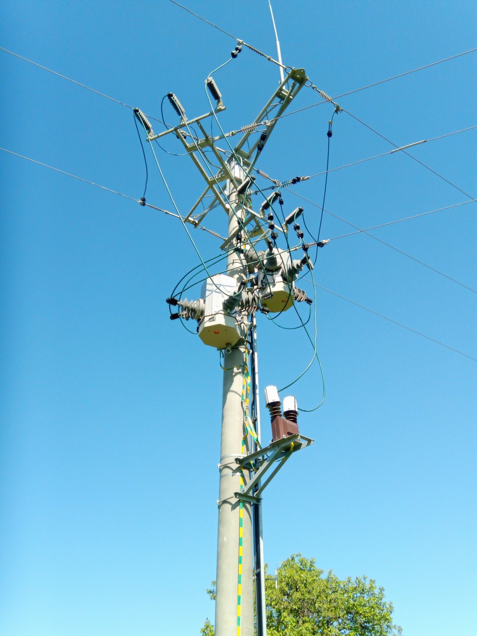 Słup energetyczny z transformatorem i izolatorami, widok z dołu na tle błękitnego nieba i fragmentu korony drzewa.
