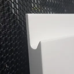 Fragment białego, lakierowanego frontu meblowego z charakterystycznym, frezowanym uchwytem, na tle czarnej siatki.