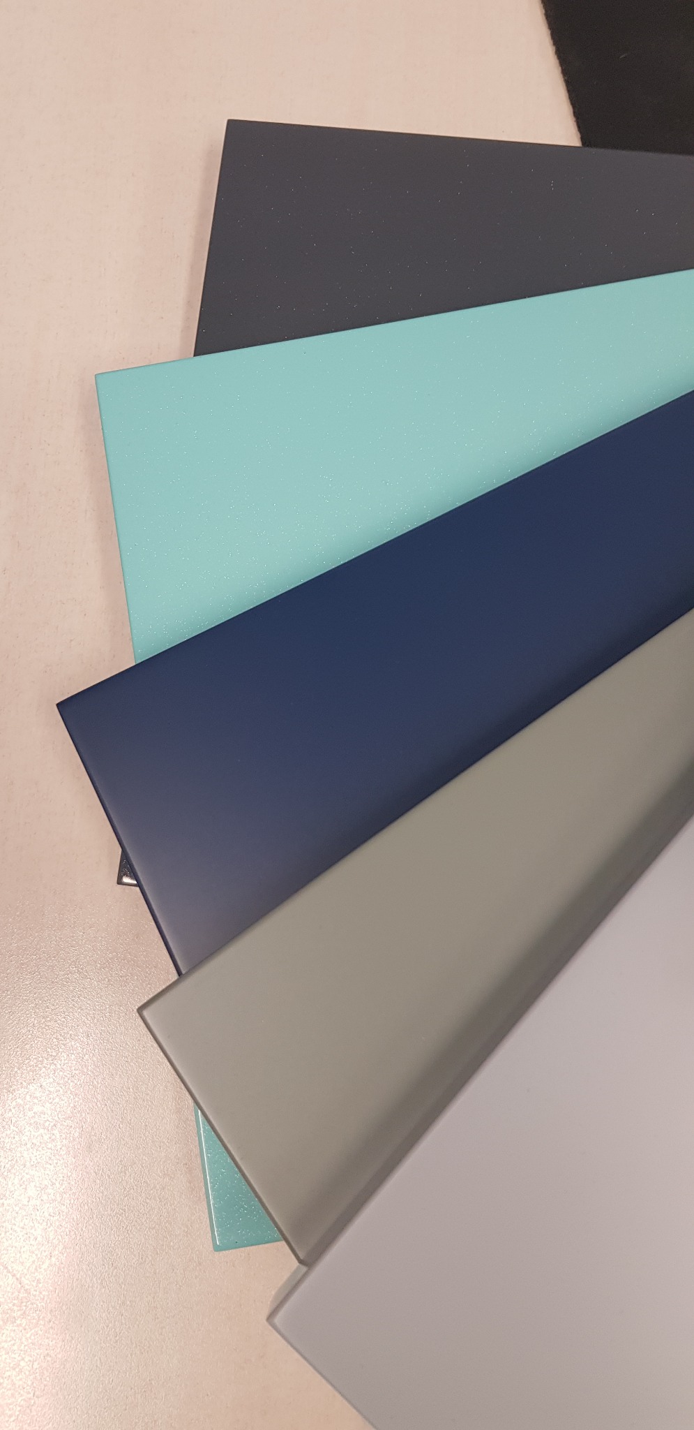 Próbki kolorów i materiałów wykończeniowych: ciemnoszary, turkusowy, granatowy, szary i biały, ułożone warstwami na jasnym blacie.