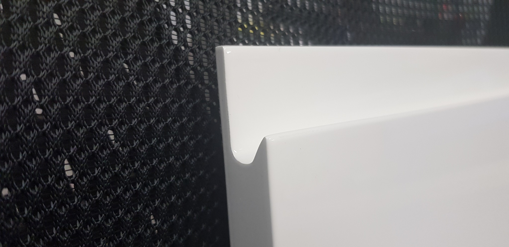 Fragment białego, lakierowanego frontu meblowego z charakterystycznym, frezowanym uchwytem, na tle czarnej siatki.