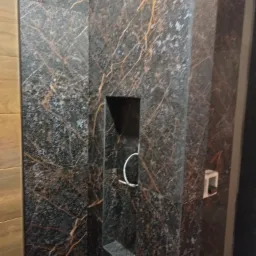 Ściana wyłożona ciemnymi płytkami imitującymi marmur z wyraźnymi złotymi żyłami, wbudowana półka w ścianie z widocznym okablowaniem, fragment drewnopodobnej okładziny po lewej stronie.