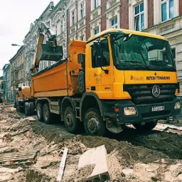 Żółta ciężarówka z łyżką koparki na tle miejskiej zabudowy, w wykopie z ziemią, widoczny element betonowy i paleta