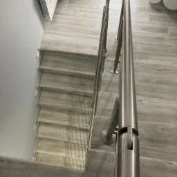 Schody wyłożone płytkami imitującymi drewno, z metalową balustradą ze stali nierdzewnej, widok z góry, szare ściany, biały grzejnik z koronkową serwetką i dwa białe balony w tle.