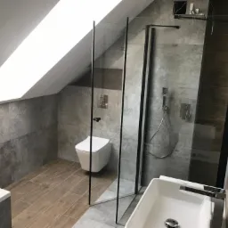 Ukończona łazienka na poddaszu z kabiną prysznicową typu walk-in, wiszącą toaletą i umywalką nablatową. Ułożone płytki imitujące drewno na podłodze i szare płytki na ścianach. Widoczne okno dachowe...