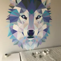 Geometryczny mural przedstawiający głowę wilka w odcieniach błękitu i fioletu, umieszczony na szarej ścianie nad zapakowanym materacem i kartonem.