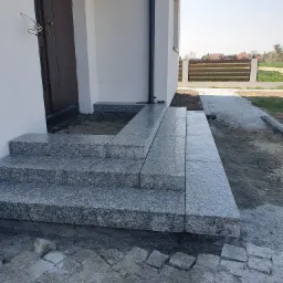 Wykończone granitowe schody wejściowe do domu jednorodzinnego z widocznymi niedokończonymi pracami brukarskimi wokół, w tle ogrodzenie panelowe i trawnik.