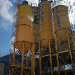 Pracownik w uprzęży linowej czyści silosy przemysłowe z napisem 'POLBET' na tle błękitnego nieba z chmurami.