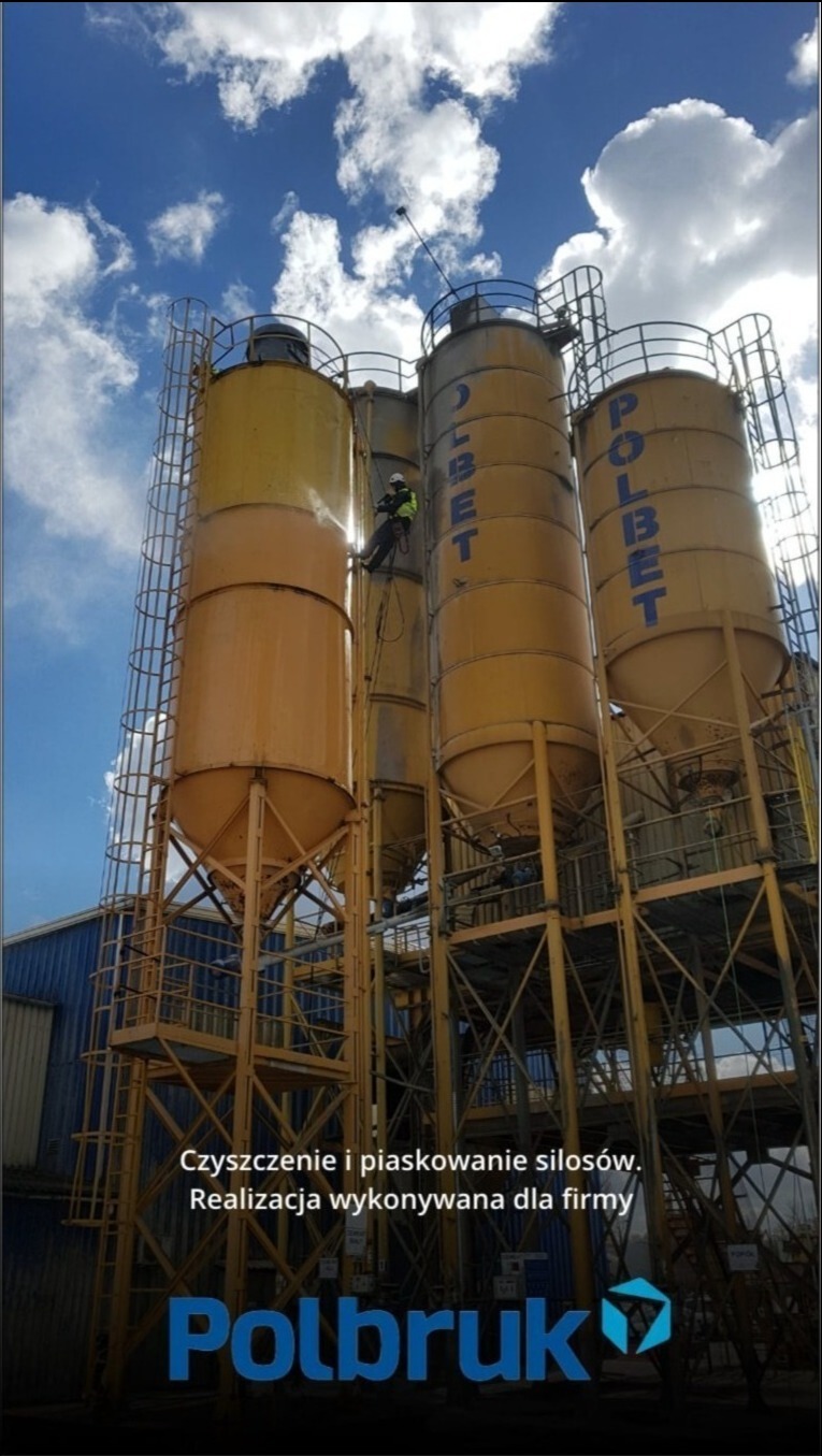 Pracownik w uprzęży linowej czyści silosy przemysłowe z napisem 'POLBET' na tle błękitnego nieba z chmurami.