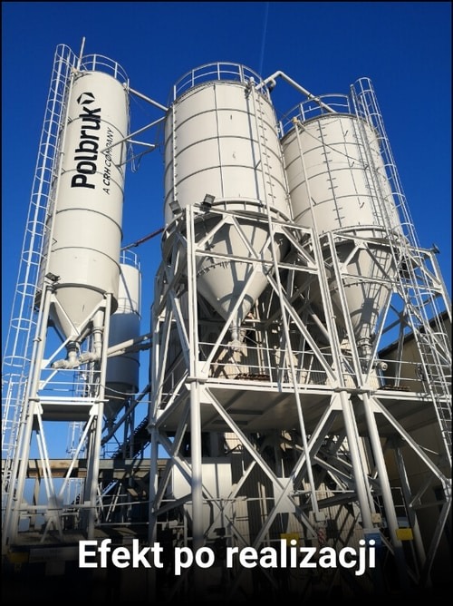 Kompleks silosów na konstrukcji stalowej, pomalowanych na biało, z logo Polbruk na jednym z nich, na tle błękitnego nieba.