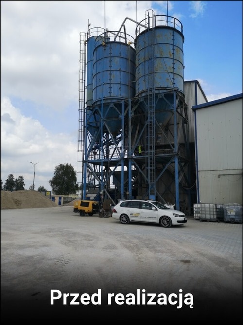 Trzy duże, niebieskie silosy przemysłowe z metalową konstrukcją wsporczą, widoczne z zewnątrz na terenie budowy; obok zaparkowany biały samochód osobowy i mała żółta koparka, a w tle budynek hali...