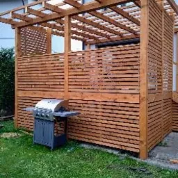 Drewniana konstrukcja z poziomych desek, prawdopodobnie altana lub zadaszenie tarasu, z grillem gazowym na zewnątrz, na tle trawnika i fragmentu budynku.
