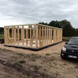 ABELIX Kamil Pawluk - Wzniesiony drewniany szkielet parterowego budynku na betonowej płycie fundamentowej, z czarną izolacją, na piaszczystym terenie z trawą, obok czarnego SUV-a.