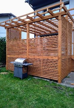 Drewniana konstrukcja z poziomych desek, prawdopodobnie altana lub zadaszenie tarasu, z grillem gazowym na zewnątrz, na tle trawnika i fragmentu budynku.