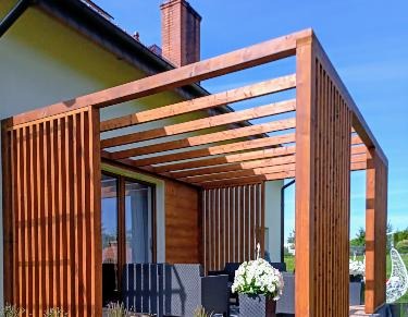 Drewniana konstrukcja zadaszenia tarasu przy domu jednorodzinnym, z widocznymi elementami dekoracyjnymi i roślinami w donicach, w słoneczny dzień.